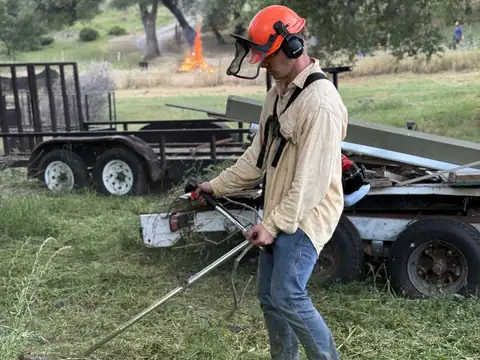 A crew member in a helmet and face shield running a brush cutter beside a trailer, a burn pile going in the field behind