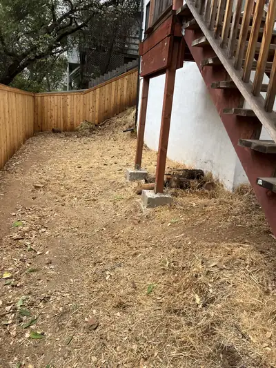 Bare, raked ground beneath a raised deck and wooden stairs, beside a wood fence