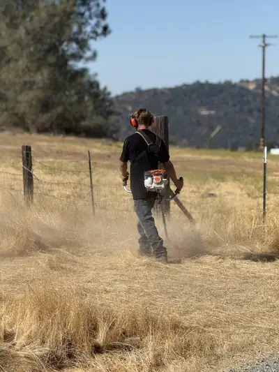 A crew member running a brush cutter through dry grass along a wire fence, dust rising, hills behind