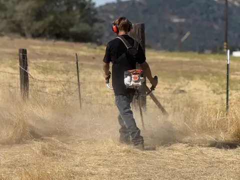 A crew member with a brush cutter working through tall dry grass beside a wire fence, hills behind