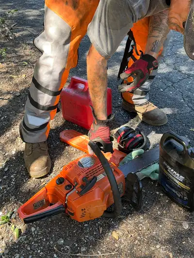 A crew member in chainsaw chaps and gloves reaching for an orange chainsaw, ear muffs and a fuel can beside it