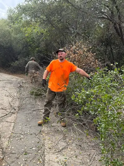 A crew member in an orange shirt and work gloves pulling cut brush off the edge of a driveway, another behind him