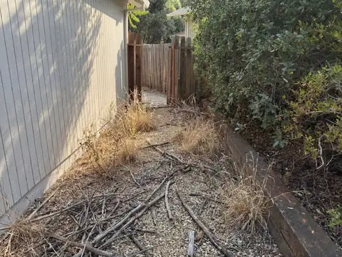 Dead branches and dry grass piled along the side of a house next to a fence