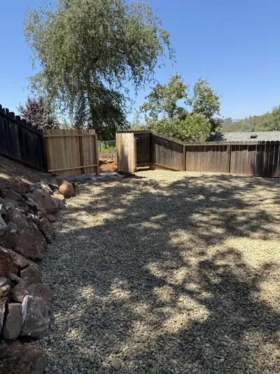 A gravel yard cleared of grass and brush along a wooden fence and a rock wall